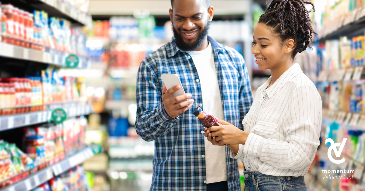 A young couple in the grocery store. The man is checking the grocery list on his cell phone while the woman is holding a bottle of sauce.