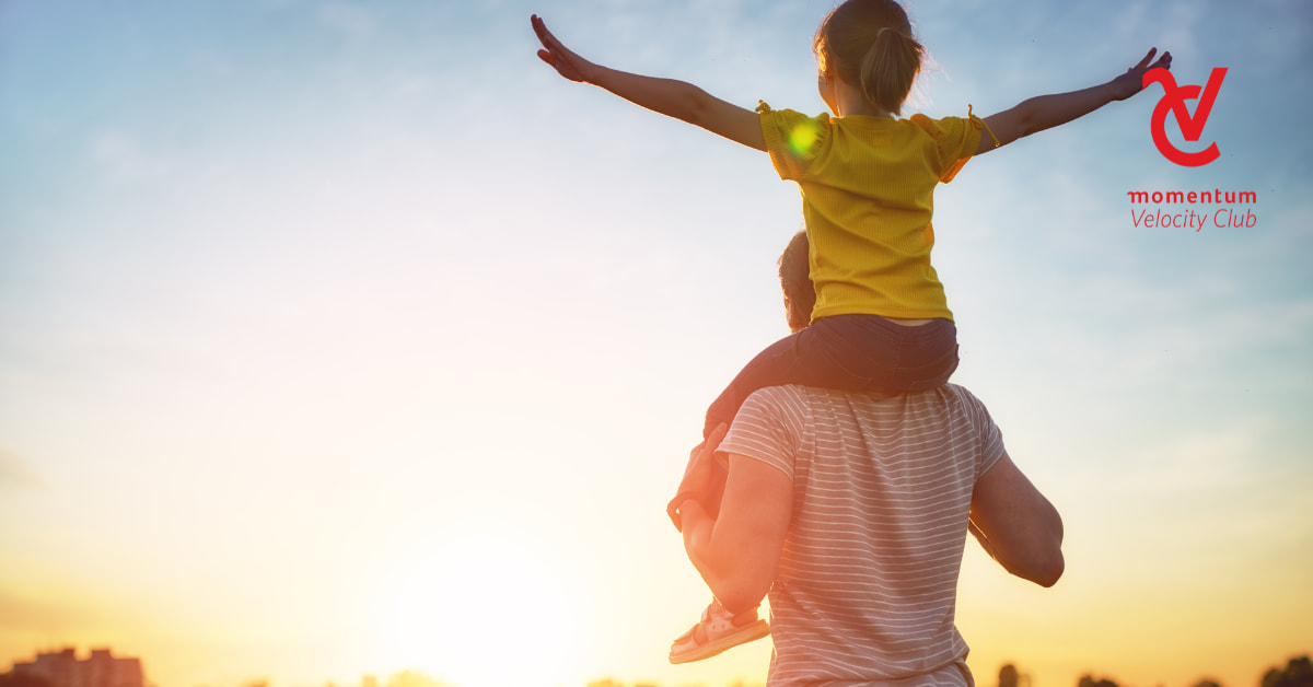 A man holding his daughter on his shoulders. The daughter's arms are stretched out as both look at the sun rising.