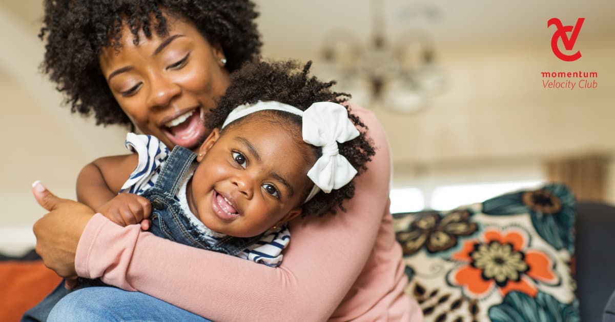 A mom is seated on a couch. She’s holding her young daughter, and they’re playing and having fun together.