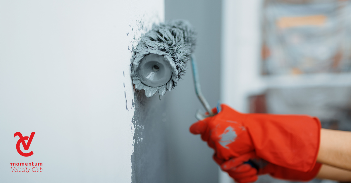 A man's hands with red gloves are holding a roller brush and painting grey paint onto a white wall.