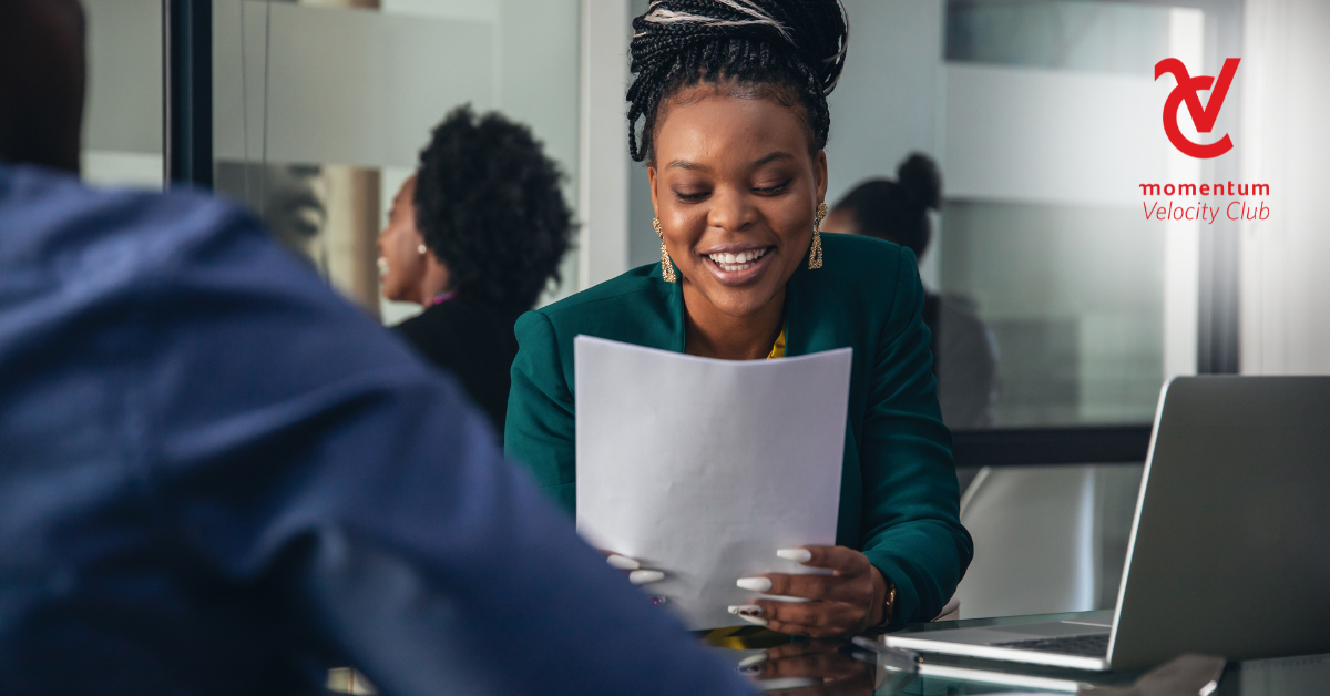 A young working professional woman smartly dressed in a green blazer is seated at her desk reviewing her retirement annuity documentation.