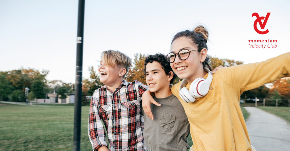 Three young children are joyfully laughing and playing together in an outdoor park, their arms wrapped around each other's shoulders.